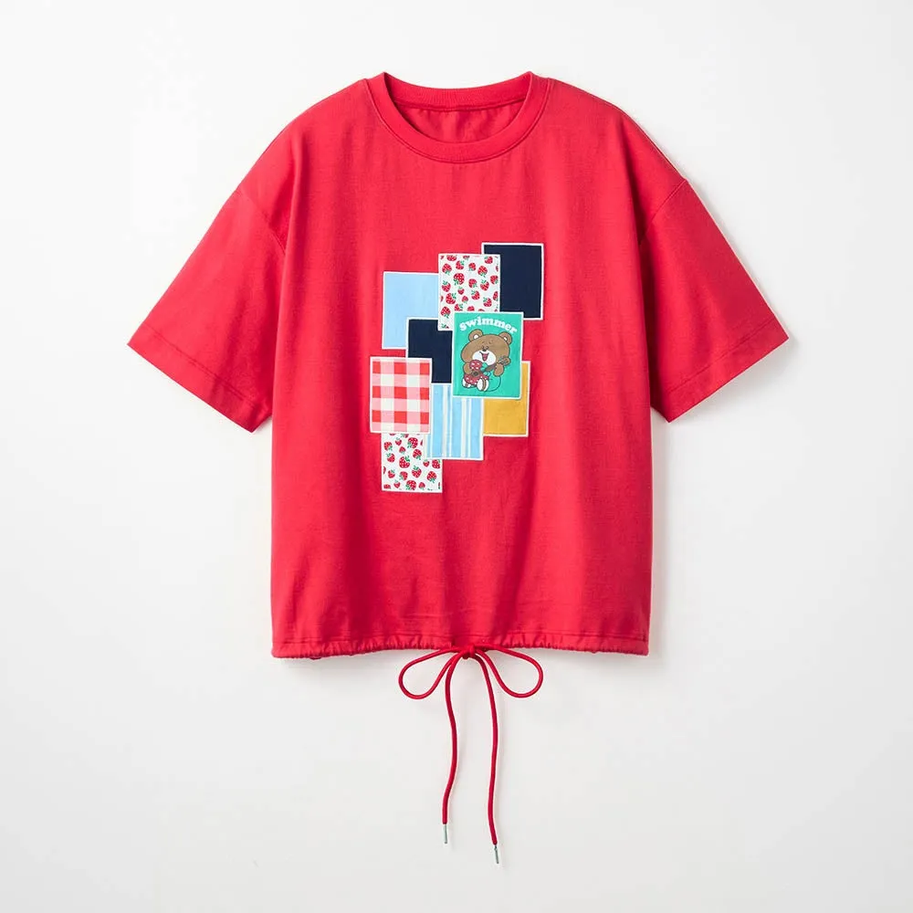 赤いTシャツ。前面には、様々な模様の四角形が組み合わされたデザインがあり、その中にクマのイラストと「swimmer」の文字が描かれている。裾には赤い紐が通されている。