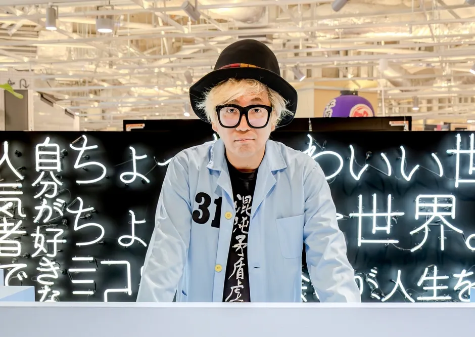 黒い帽子と大きなメガネをかけた男性が、ネオンサインの前で立っている写真。