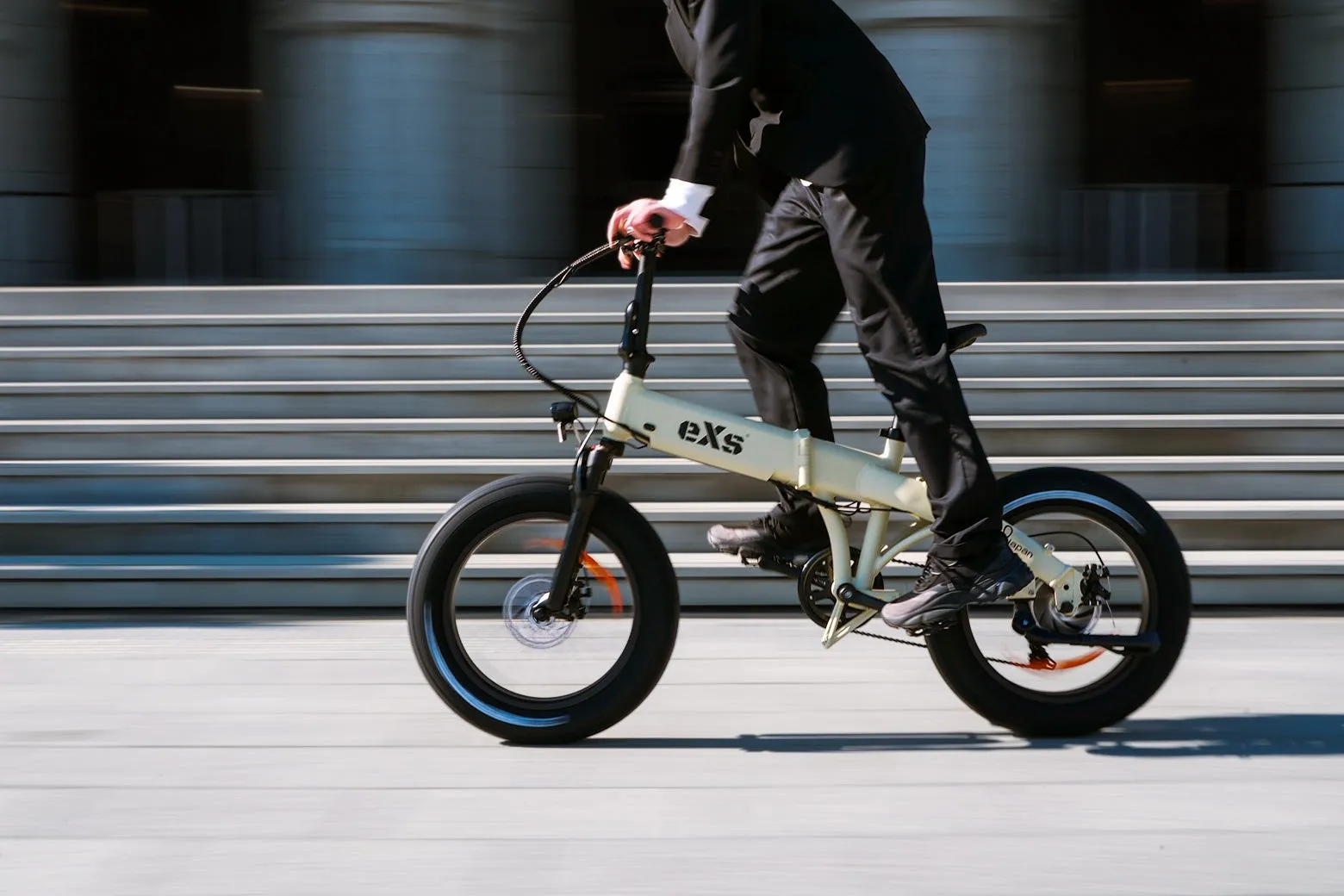スーツを着た男性がeXsの電動自転車に乗っている写真。自転車は白く、背景には階段がある。