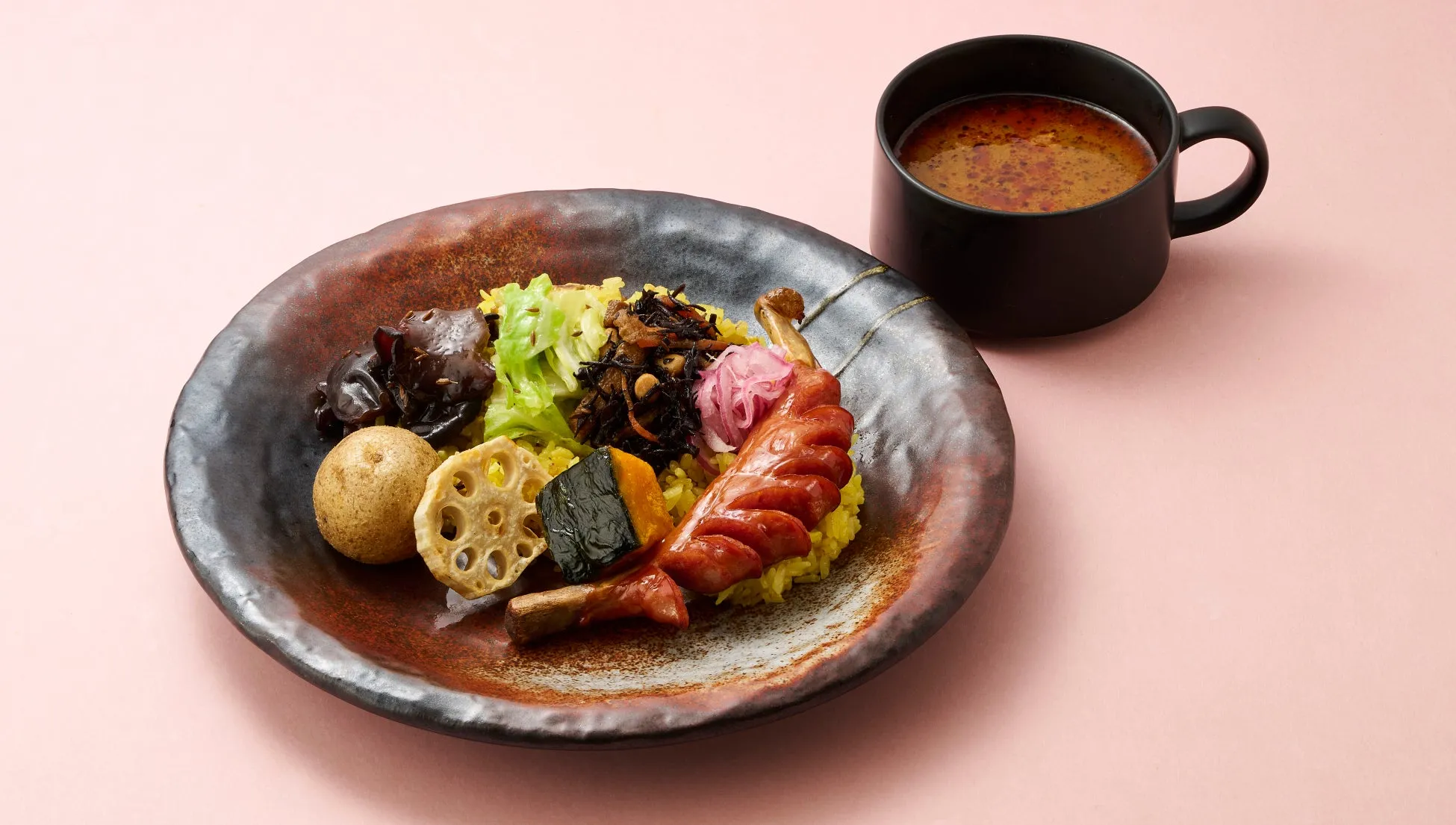 カレーライスのような料理が、黒いカップに入ったスープと一緒に、茶色い皿に盛られています。ピンク色の背景です。