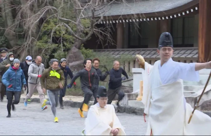 男性たちが走っている様子を捉えた画像です。手前には白い衣装をまとった人物がおり、その後ろを多くの人が追いかけるように走っています。背景には建物や木々が見え、屋外でのイベントのようです。