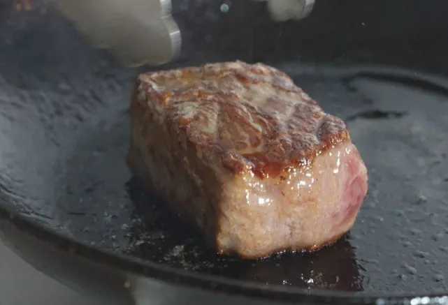 ステーキがフライパンで焼かれている様子。火加減を見ている様子が写っている。