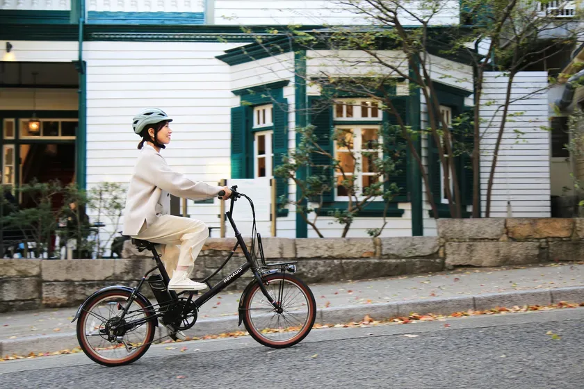 女性が自転車に乗っている写真。自転車は黒色で、女性はベージュ色の服を着ており、ヘルメットを被っている。背景には白い壁と緑色の窓枠の建物がある。