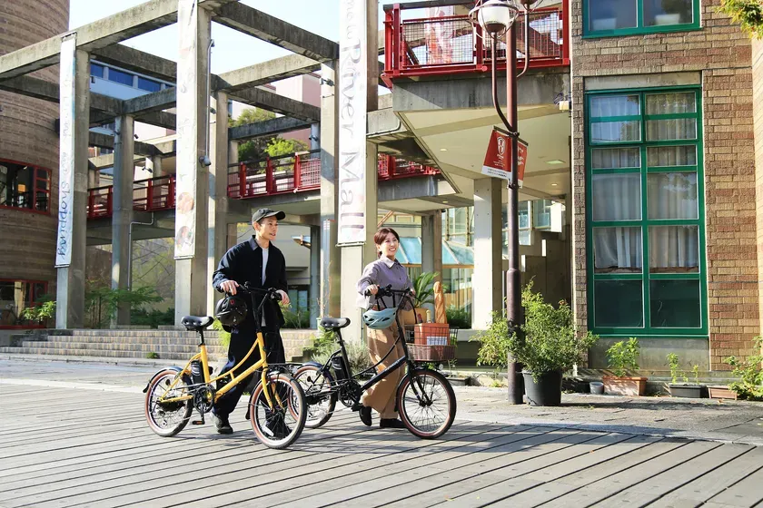 二人の男女が自転車を押して歩いている様子が写っています。背景にはショッピングモールの建物があり、日差しの良い屋外での活動を示唆しています。