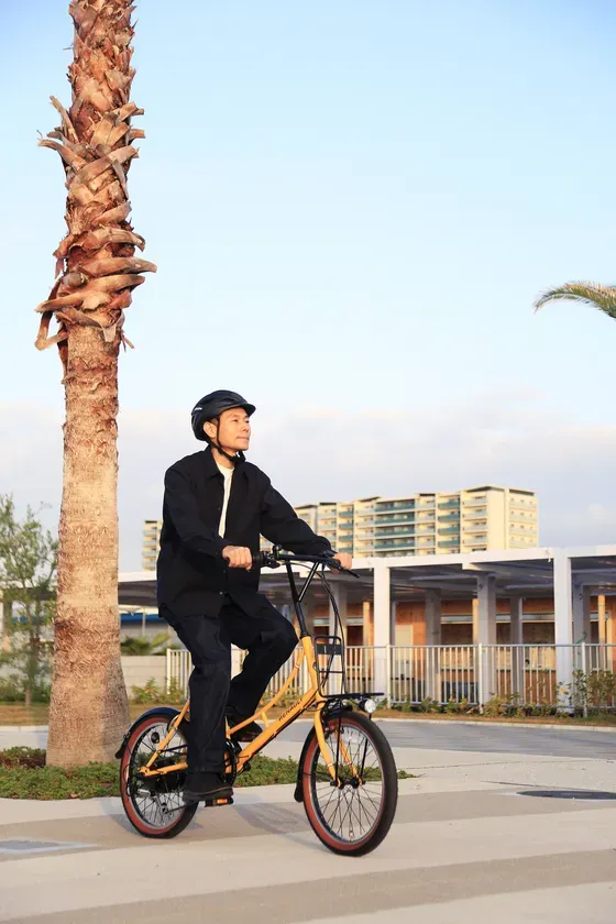 自転車に乗っている男性の写真。彼は黒い服を着ており、ヘルメットを着用している。自転車は黄色で、背景には建物と木々がある。