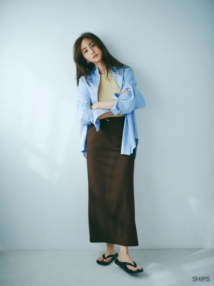 女性が青いシャツと茶色のスカートを着て、白い壁に寄りかかっています。