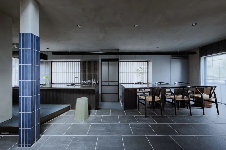 モダンなキッチンとダイニングエリア。濃い色の木材と暗い色の床、青いタイル張りの柱が特徴的。洗練されたデザインで、モダンでありながら落ち着いた雰囲気。