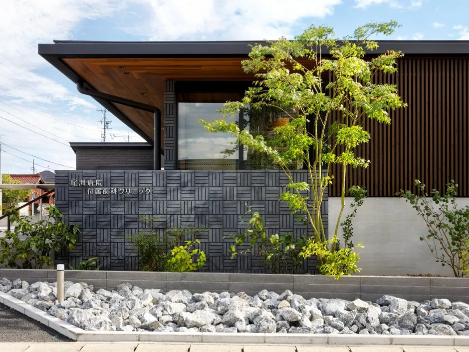 モダンなデザインの歯科医院の外観。グレーのブロック塀、木材、植栽が組み合わさり、洗練された印象を与えます。建物の正面には庭があり、白い石が敷き詰められています。