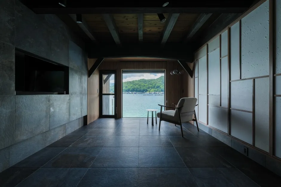 窓から海の見えるモダンなリビングルーム。グレーのタイル張りの床と壁、木の梁とパネル、アームチェアと小さなテーブルが配置され、洗練された空間を演出している。
