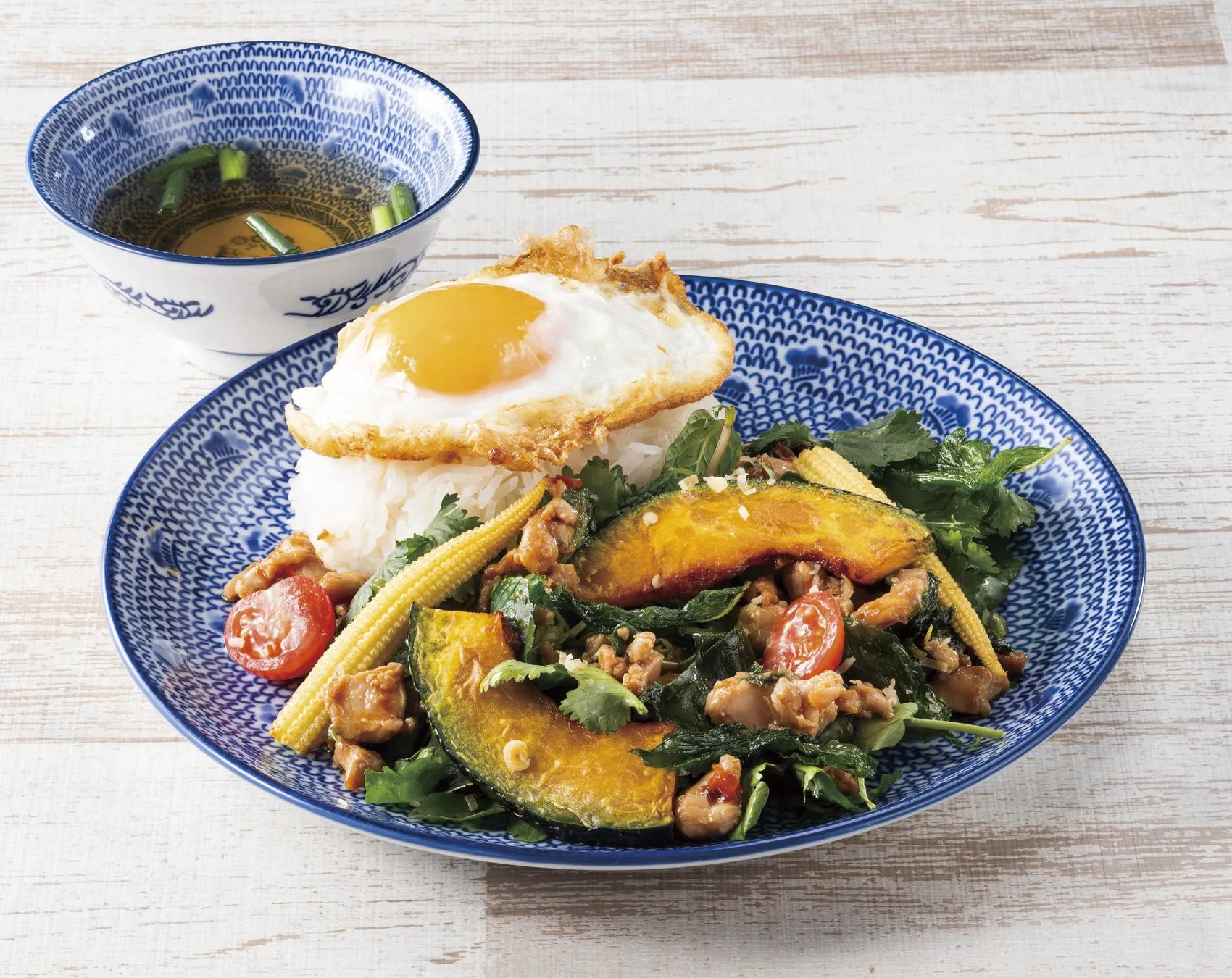 白いご飯の上に目玉焼きが乗っており、その周りにカボチャ、鶏肉、ミニトマト、コーン、ハーブなどが盛られた料理。手前にはスープが入った器が置かれている。