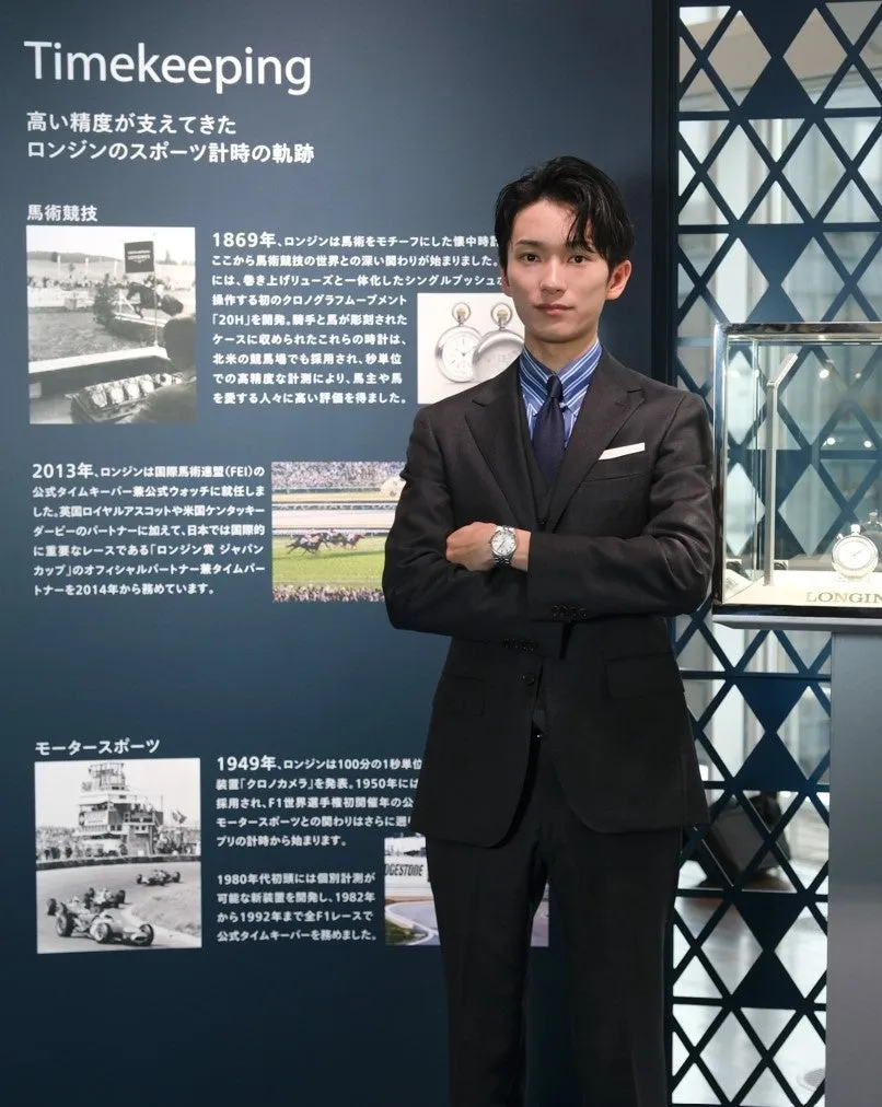 ロンジンの時計の展示会で、スーツを着た男性が写っている写真。背景には、ロンジンの歴史とスポーツ計時の軌跡について説明するパネルがあり、時計やモータースポーツ、馬術競技などに関する情報が掲載されている。