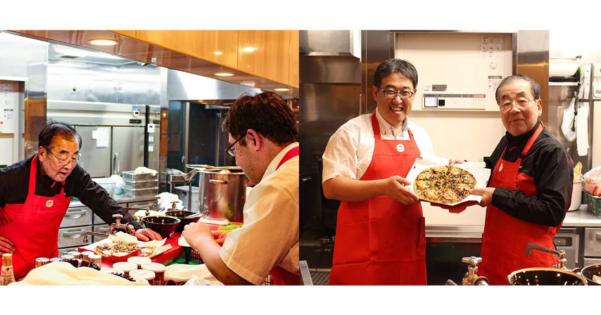 2人の男性が厨房でピザを調理している様子が写っています。一人はピザを手に持ち、もう一人は調理台で作業をしています。