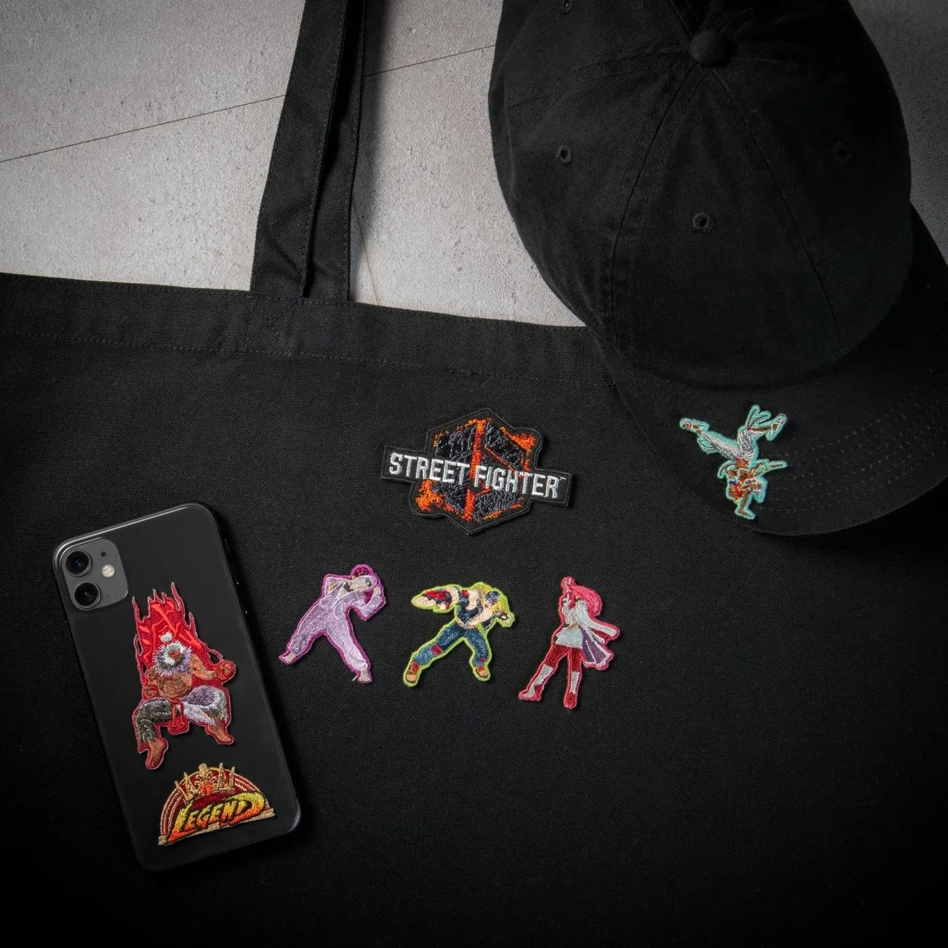 黒いトートバッグ、帽子、スマートフォン、ワッペンが写っている画像。バッグには「STREET FIGHTER」のワッペンと、キャラクターのワッペン、スマートフォンにはキャラクターのワッペンと「LEGEND」の文字が見える。