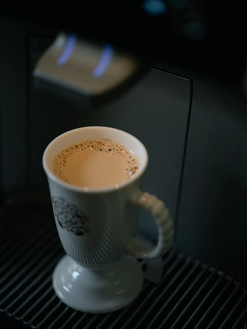 白いマグカップに入ったコーヒー。マグカップは、コーヒーメーカーの金属製の棚の上に置かれています。