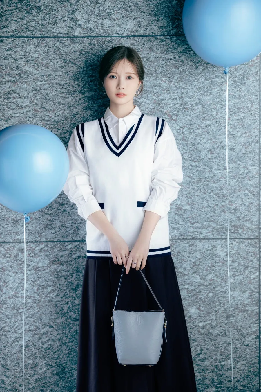 女性が白いシャツとベスト、黒いスカートを着て青い風船のそばに立つ着用イメージ