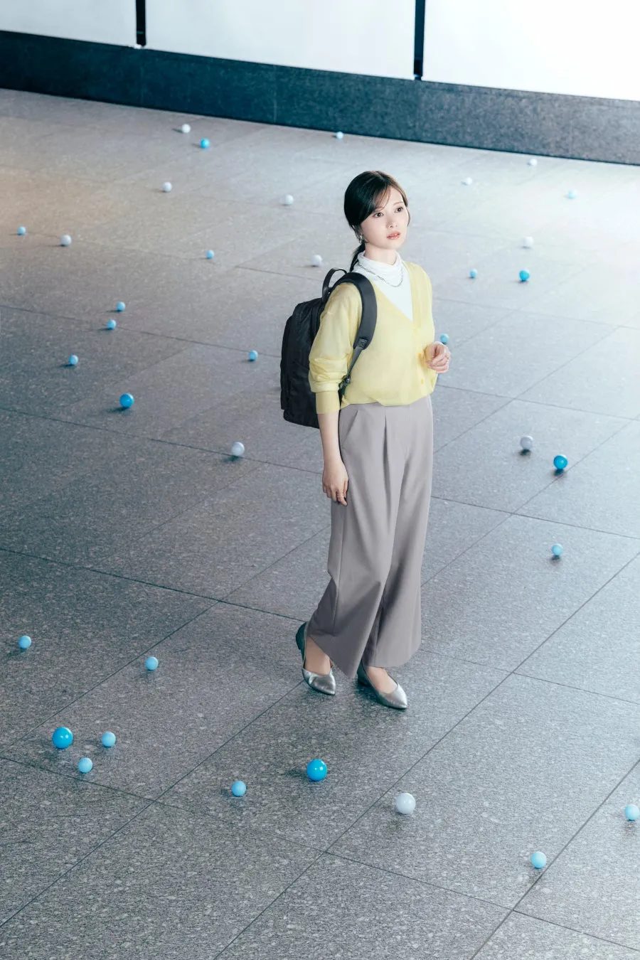 女性がカーディガン、パンツ、リュックサックを身につけ、青いボールを見つめる着用イメージ