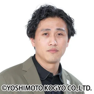 男性が正面を向いて立っている写真です。彼は黒いシャツとベージュのジャケットを着ています。彼の髪は黒く、カールしています。背景は白です。