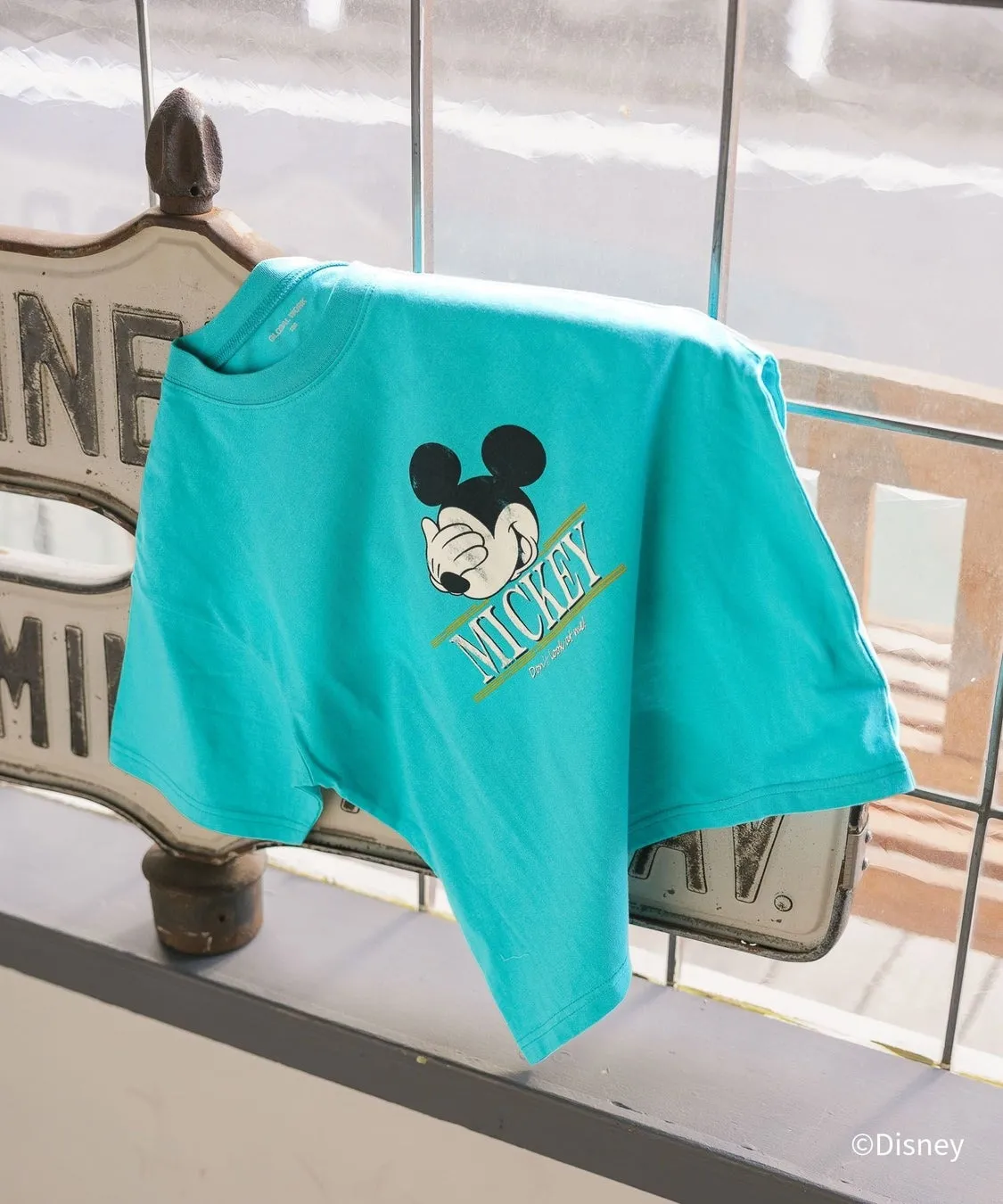 ミッキーマウスの顔がプリントされた水色のTシャツが窓枠に掛けられている写真です。Tシャツの左側には、古い看板の一部が見えます。背景は窓と外の景色です。