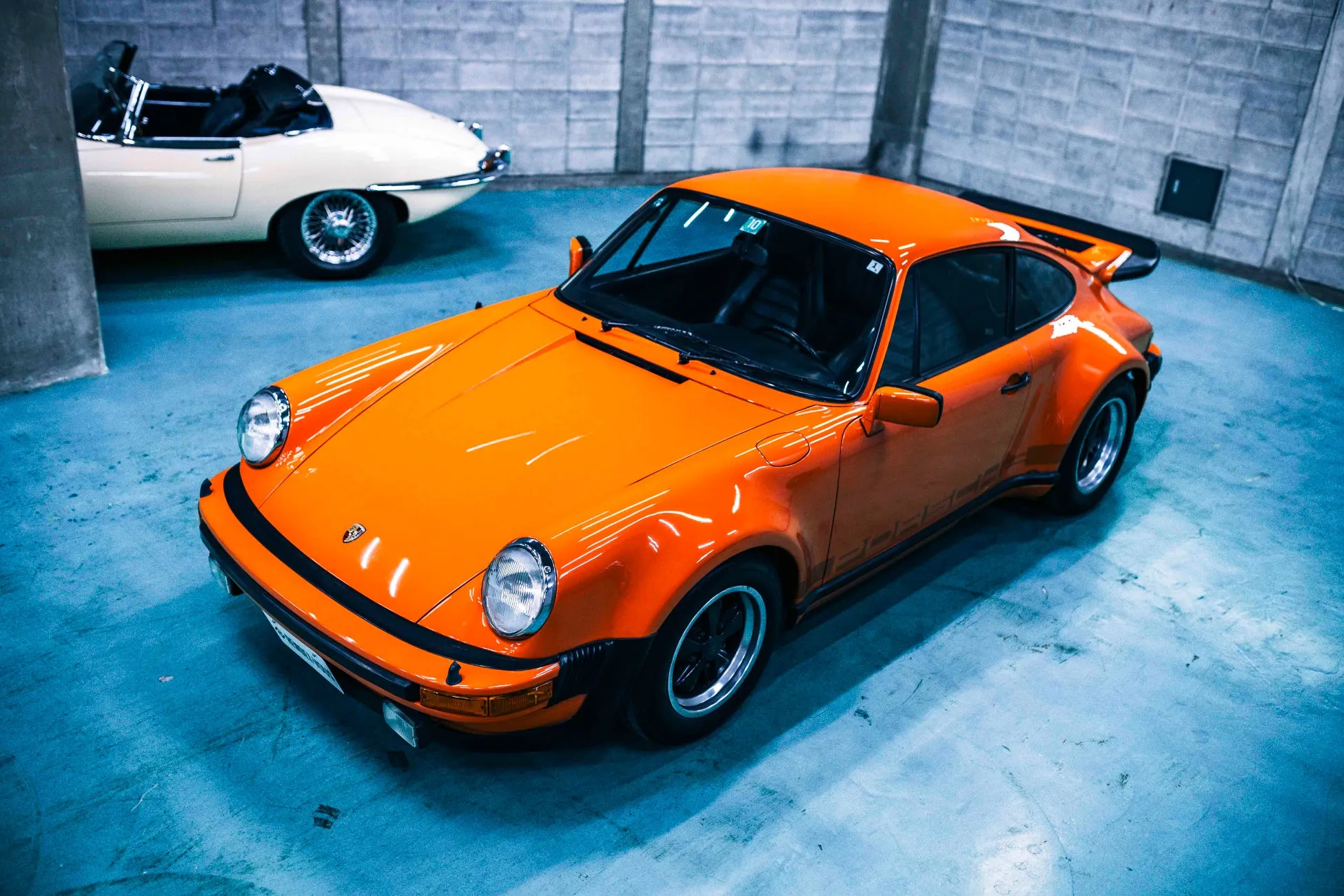 オレンジ色のポルシェと白い旧車が写っている写真。ポルシェは駐車場に停まっており、もう一台の車は背景にぼやけて見える。