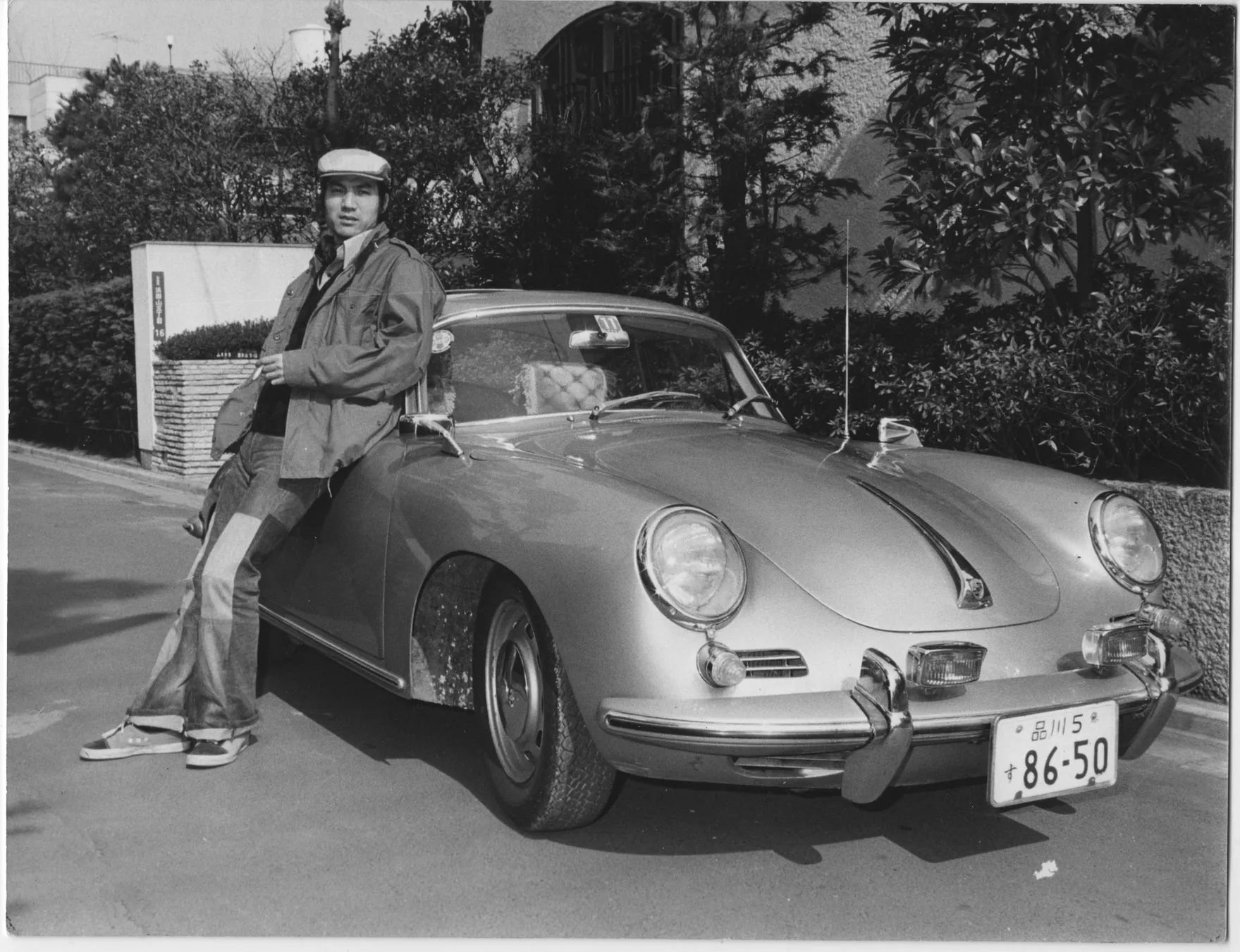 男性がポルシェに寄りかかっているモノクロ写真。男性は帽子とジャケットを着て、車に寄りかかっており、背景には木々がある。