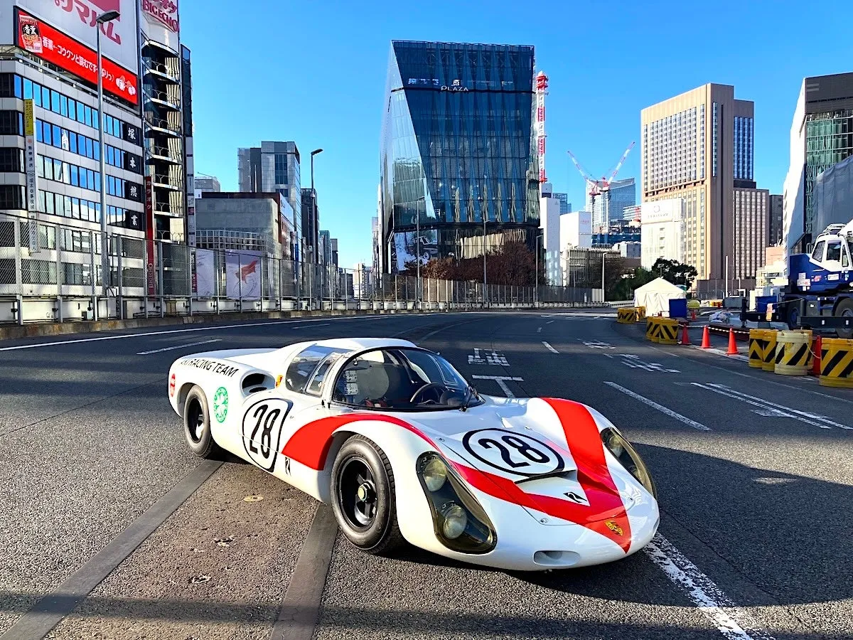晴れた日の都会の道路に停まっているレーシングカーの写真です。車は白と赤のカラーリングで、ゼッケン28が大きく表示されています。周囲には高層ビルが立ち並び、建設中のクレーンも見えます。