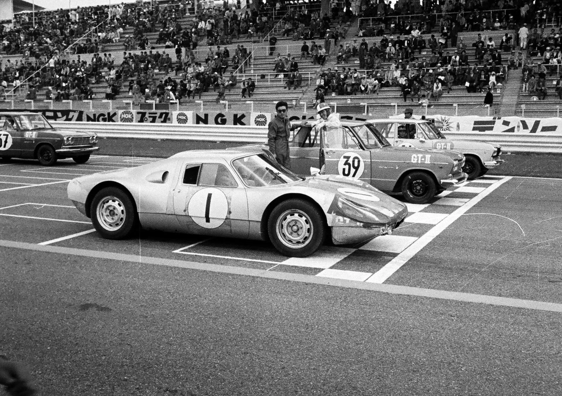 レースカーがスタートラインに並んでいるモノクロ写真です。観客席には多くの観客が座っています。