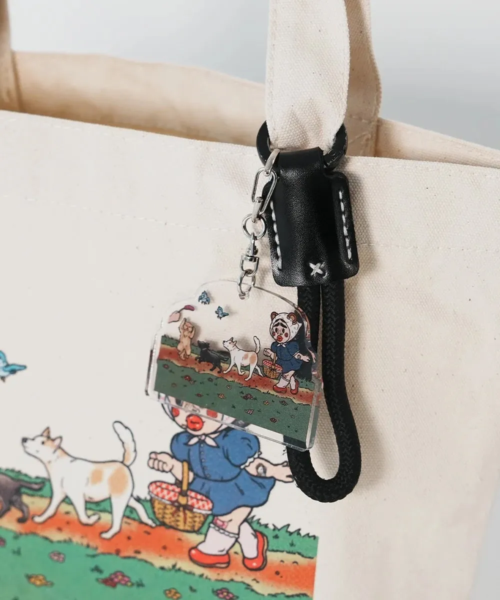 白いトートバッグに、犬と少女が描かれたキーホルダーが付いている写真。キーホルダーには、犬と少女が草むらを歩く様子が描かれている。