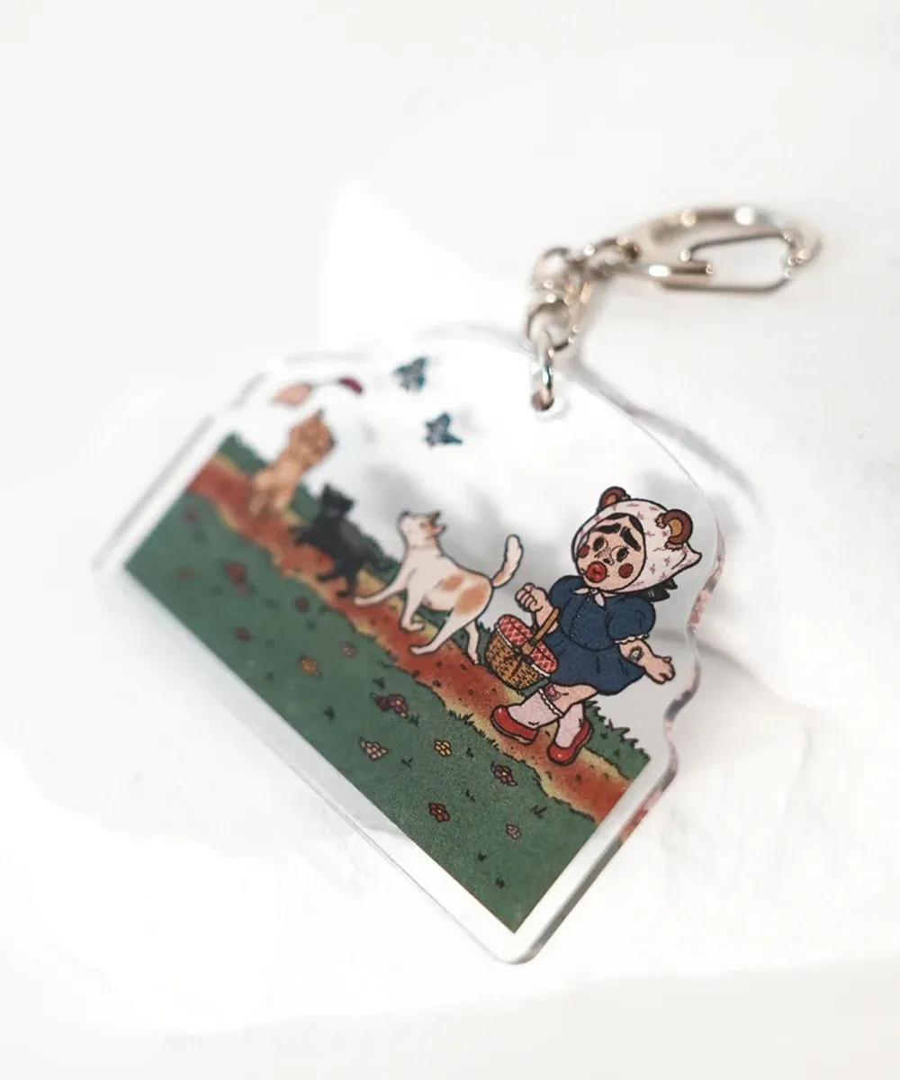 クマの帽子をかぶった女の子が、バスケットを持って猫と一緒に歩いているキーホルダー。