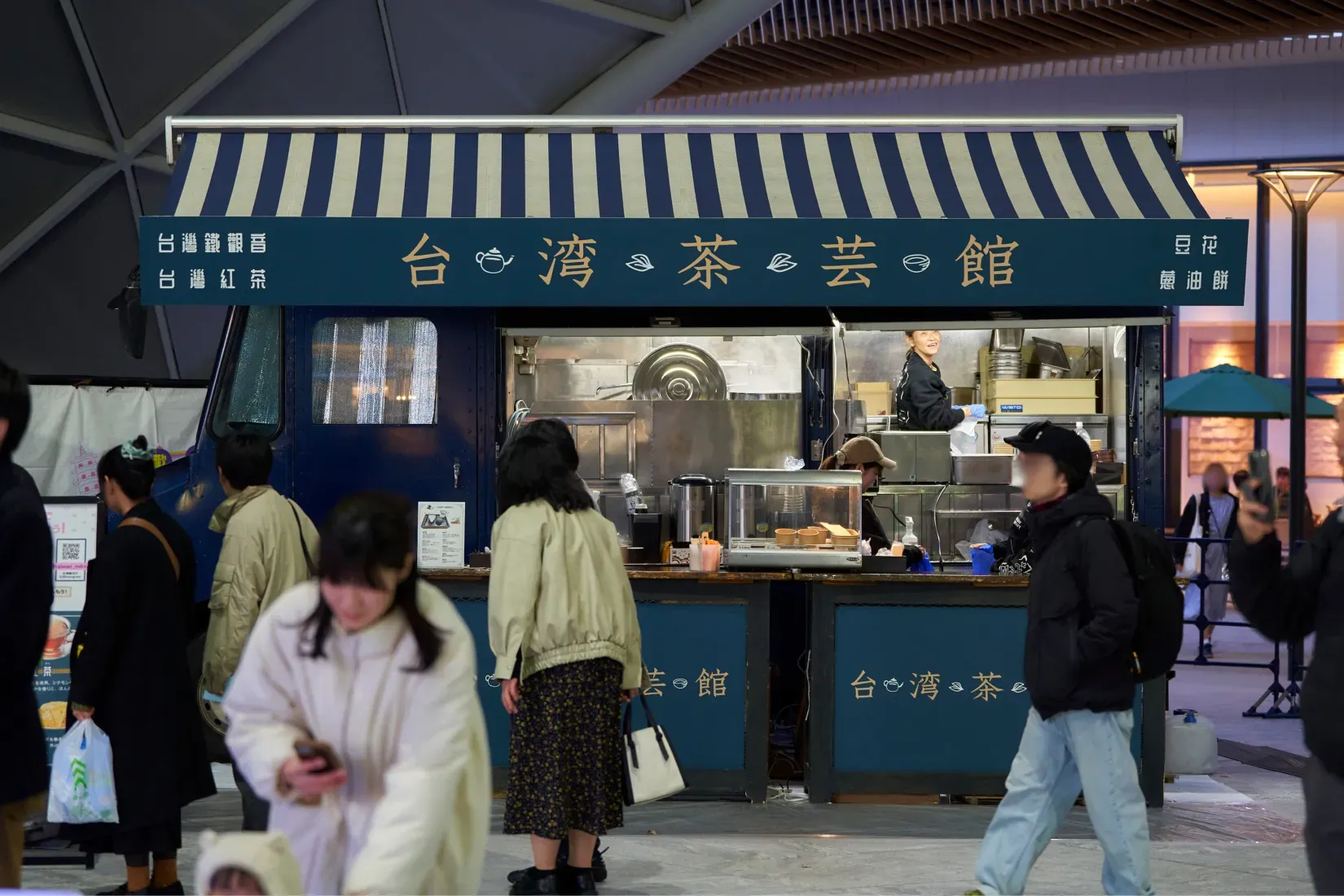 台湾茶芸館の屋台の様子を写した写真。屋台の看板には台湾のお茶の種類と、メニューの豆花と葱油餅の文字が見える。店員と客の姿も捉えられている。