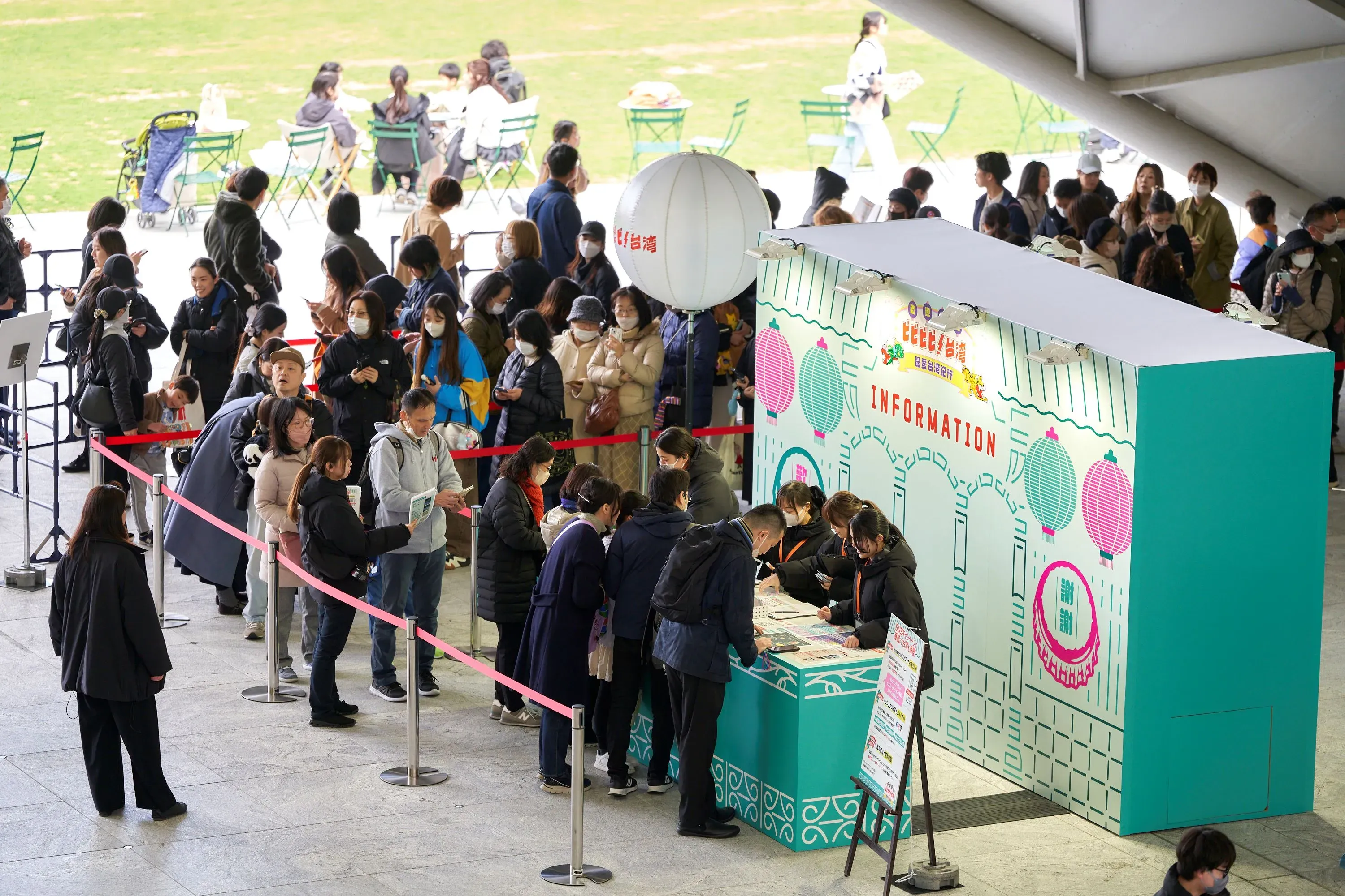 イベント会場のインフォメーションブースの前に並ぶ人々の様子を捉えた写真です。人々はマスクを着用しており、イベントに参加するために集まっているようです。