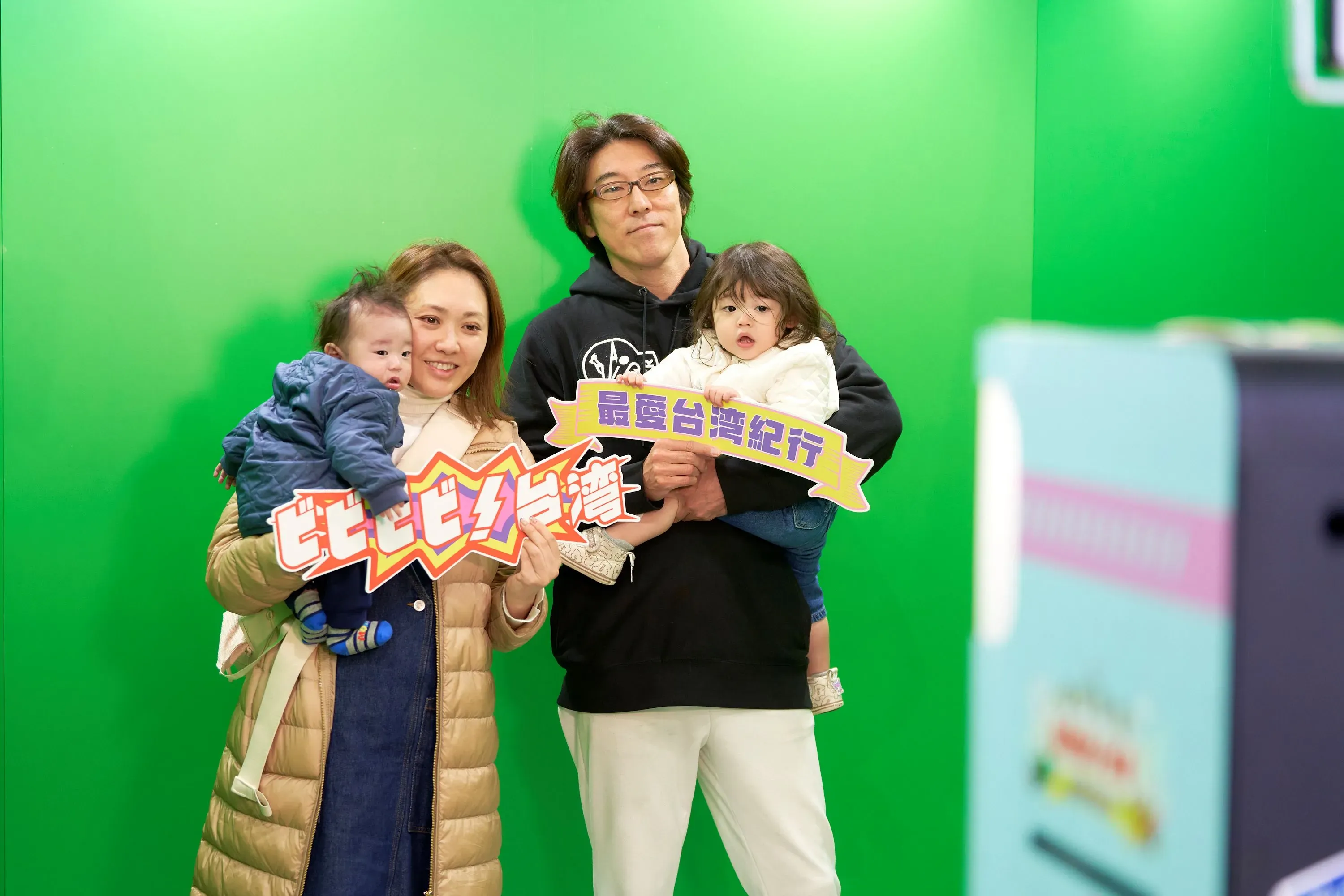 家族4人が写った写真。両親がそれぞれ子供を抱っこしており、子供たちはそれぞれ看板を持っている。背景は緑色。