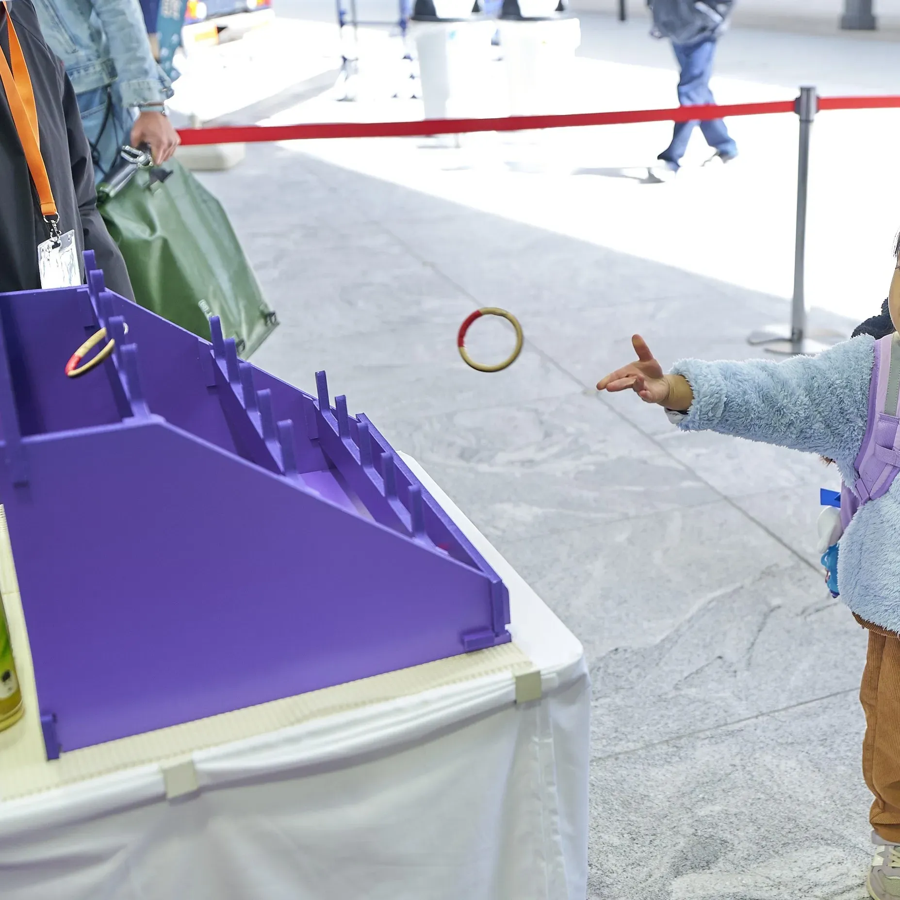 輪投げをしている子供の姿を捉えた写真です。子供は、紫色の輪投げ台に向かって輪を投げています。背景には、他の人々や赤いロープが見えます。