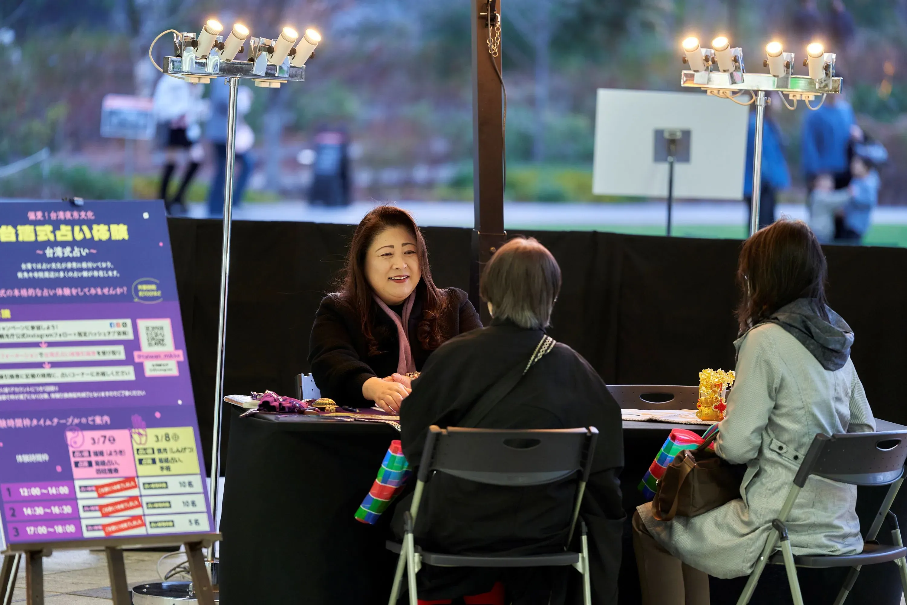 台湾式占いのイベントの様子を写した写真です。女性占い師が2人のお客様と対面しており、テーブルには占いの道具や資料が置かれています。背景にはイベントの看板や照明が見えます。