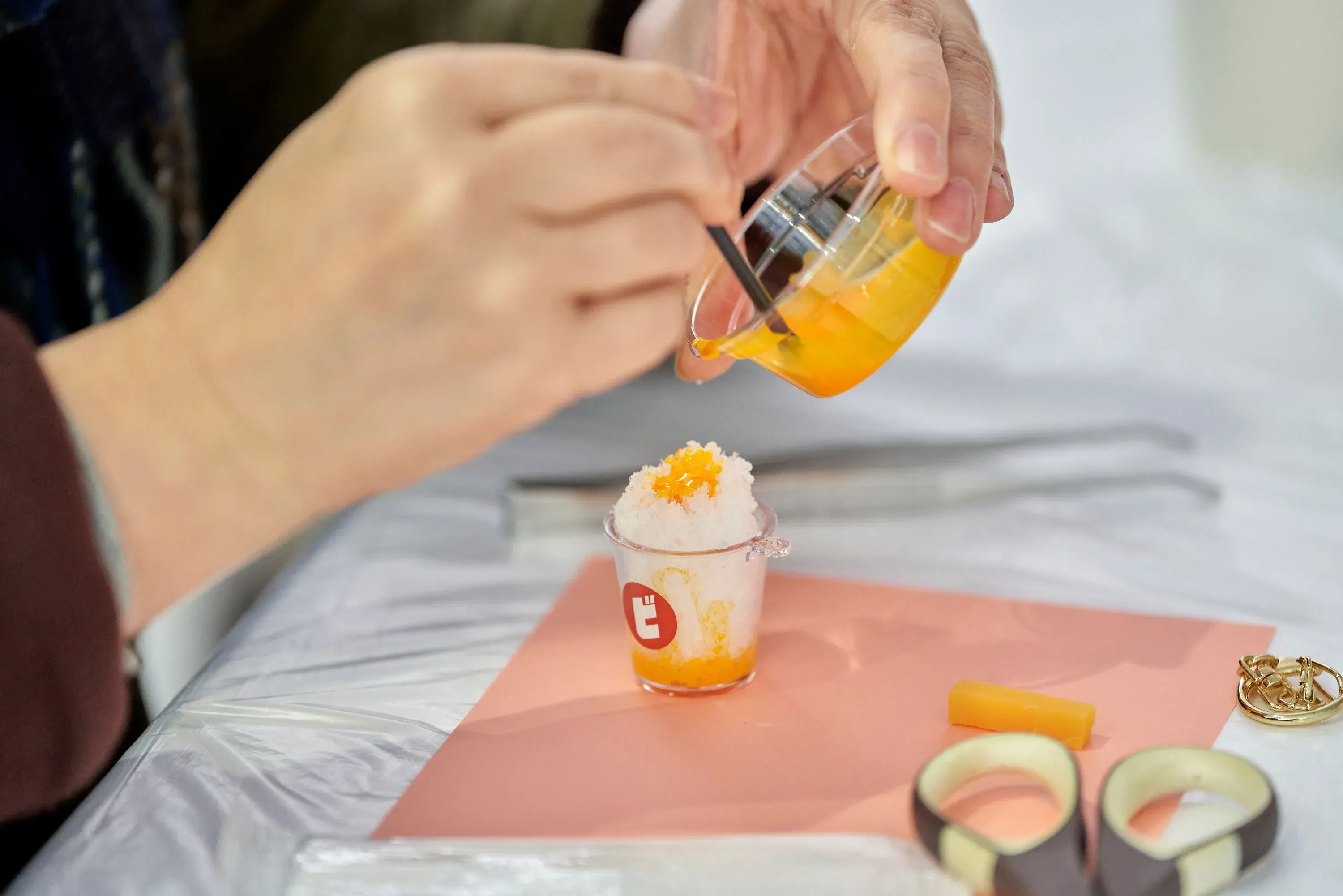 透明な容器から、かき氷にシロップをかけている様子を写した写真です。かき氷は透明なカップに入っており、すでにシロップがかけられています。周囲には、手、カッター、アクセサリー、その他の材料が見えます。