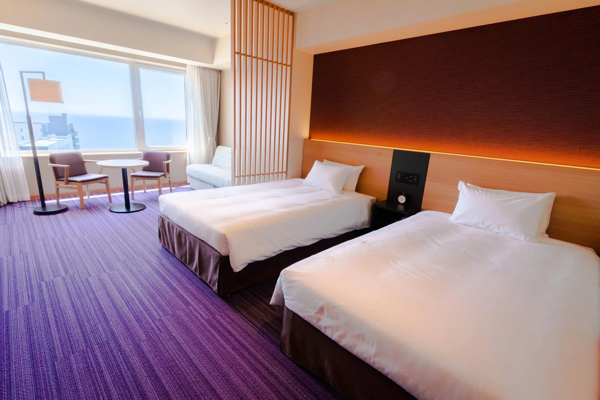 2つのベッドとテーブルと椅子のあるホテルの部屋。窓からは海が見え、明るい光が差し込んでいる。