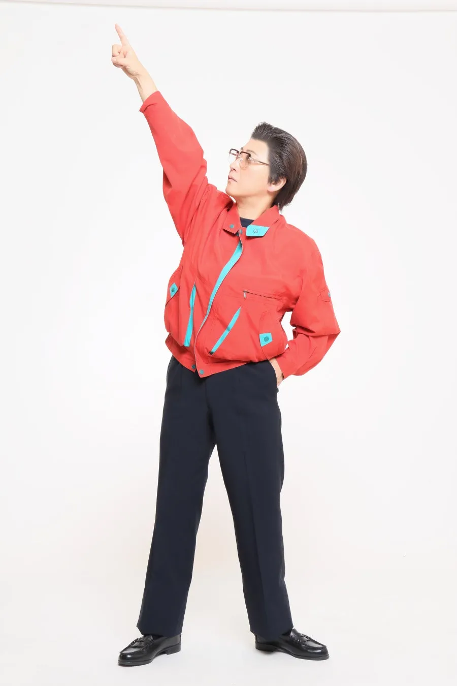 赤いジャケットと黒いズボンを着て、指をさしている男性の全身写真。彼は眼鏡をかけており、白背景に立っている。