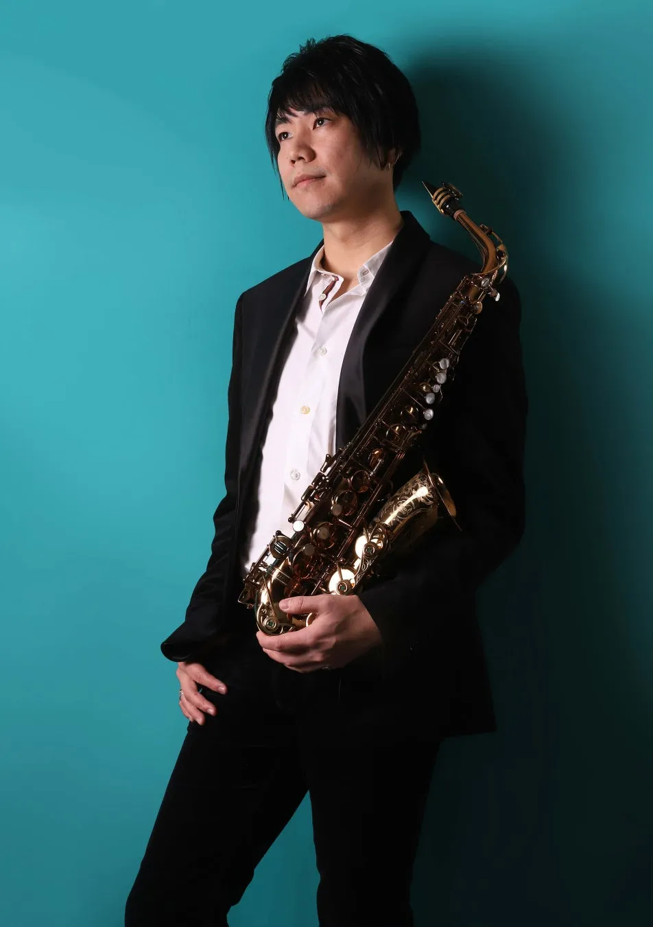 サックスを抱えた男性が、青色の背景をバックに立っている写真です。