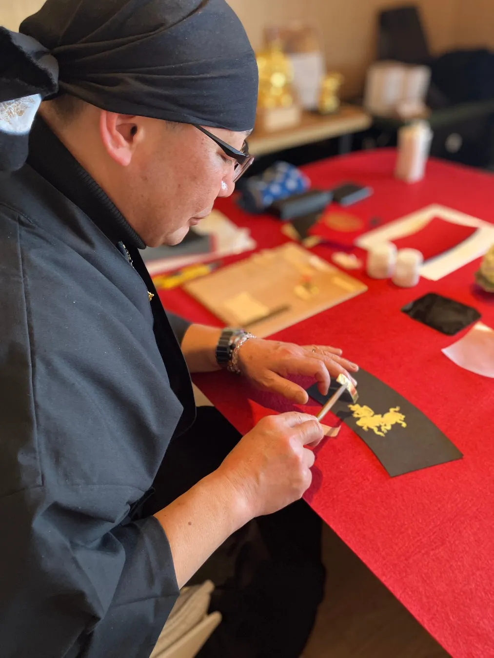 職人が、赤いテーブルの上で、金色の装飾が施された黒い扇子のようなものに作業をしている様子。