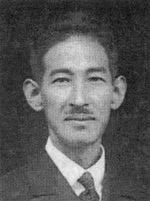 男性のポートレート写真。黒いスーツにネクタイ姿で、穏やかな表情をしている。