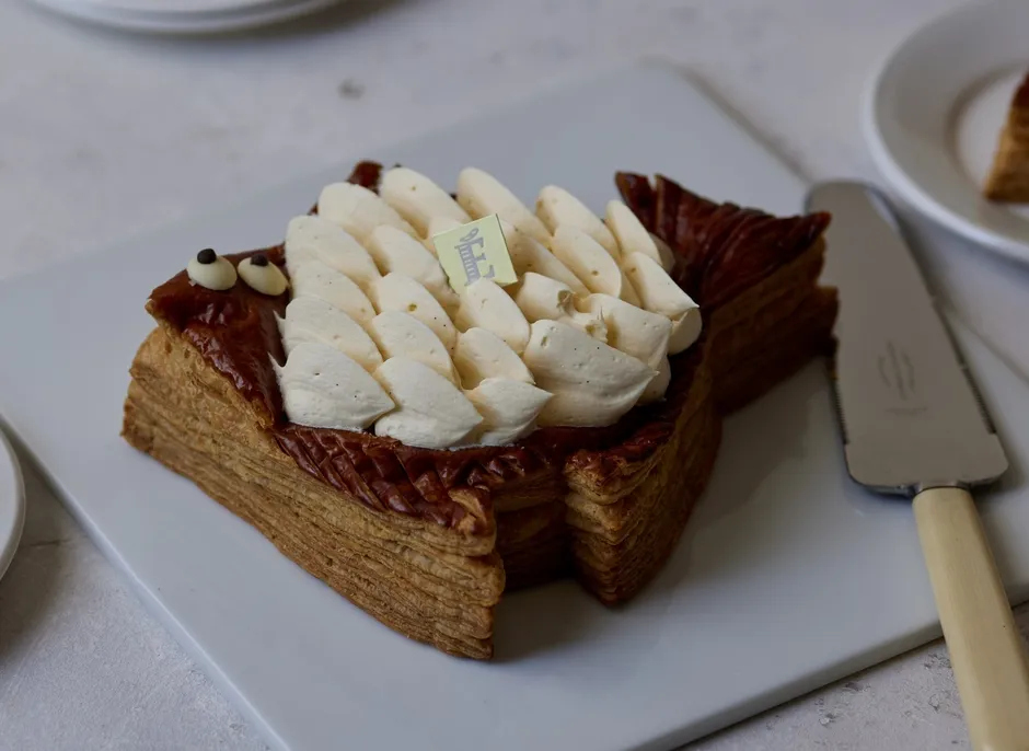 魚の形をしたケーキが白い皿に乗っています。ケーキの上には白いクリームがトッピングされ、チョコレートの目がついています。ケーキの側面には、層状のペストリーが見えます。ケーキの横にはケーキサーバーが置かれています。