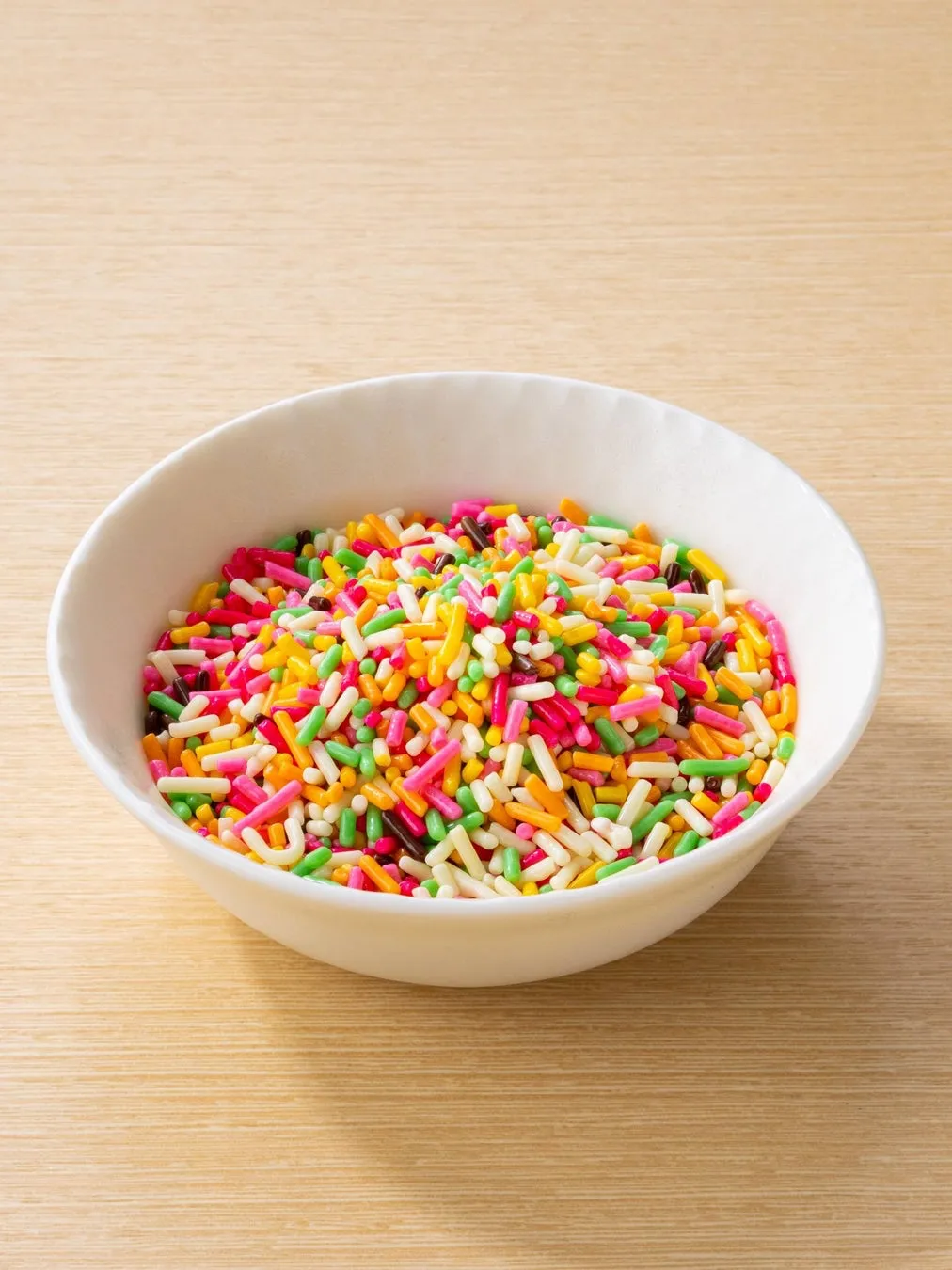白い器に入ったカラフルなトッピングの画像です。様々な色の細長いスプリンクルが器いっぱいに詰められています。背景は木目調のテーブルで、シンプルながらもカラフルなコントラストが目を引きます。