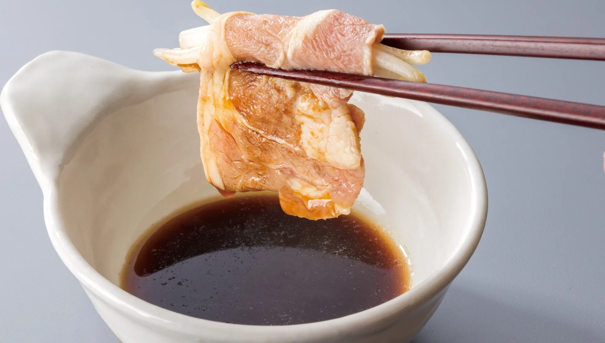 箸で持たれた豚肉とネギの巻き物と、タレが入った器のクローズアップ写真。豚肉は薄切りで、ネギを包み込んでいる。器は白く、タレは濃い茶色をしている。