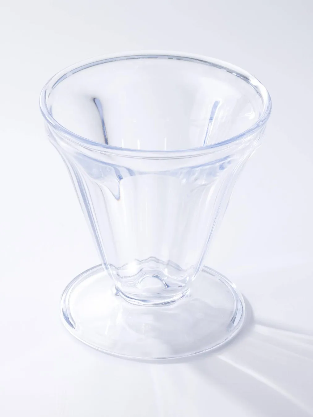 透明なパフェグラスのクローズアップ写真。グラスは、脚と広がった縁のある、背の高い円錐形をしている。グラスは平らな白い表面に置かれており、影が写っている。