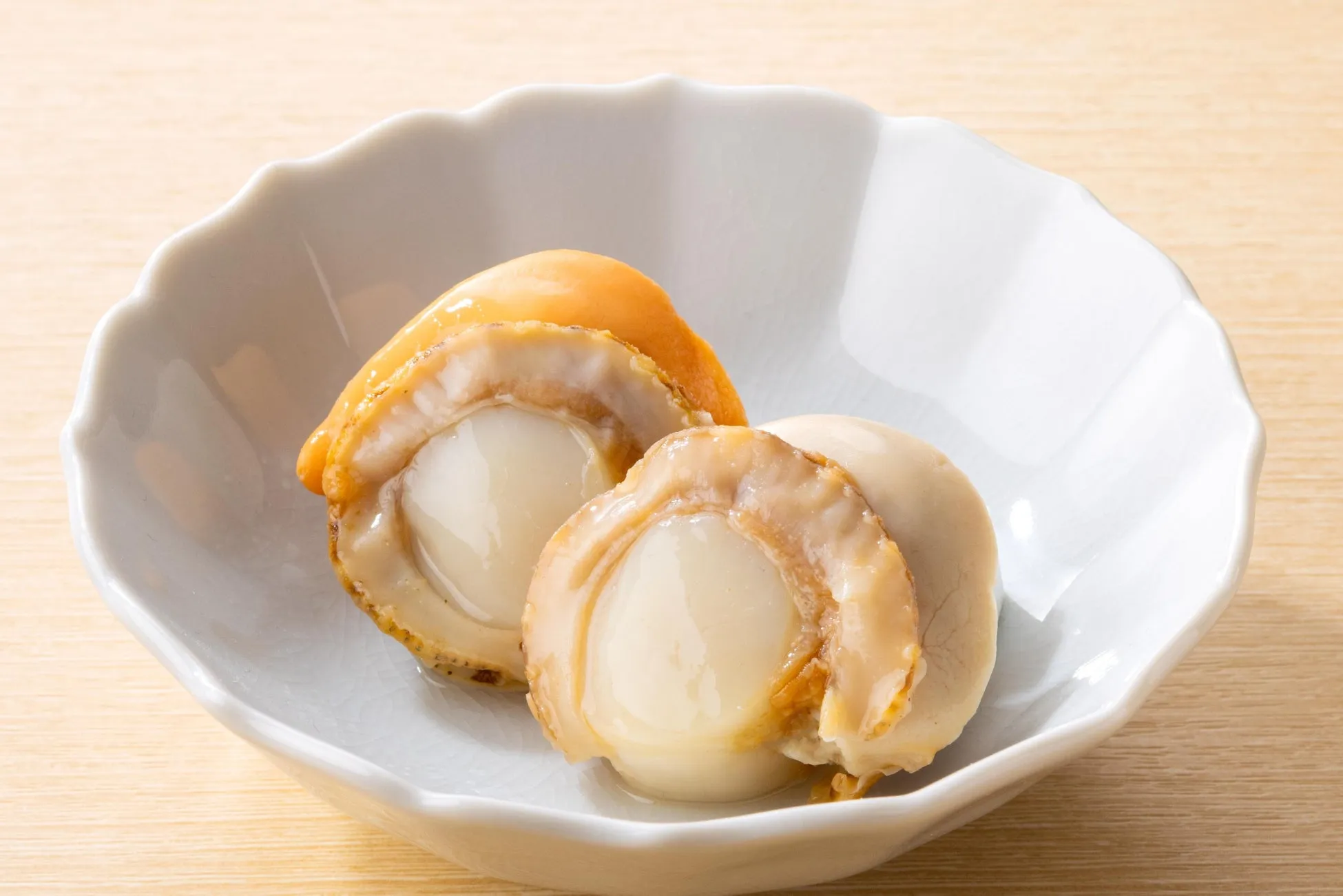 お皿に盛り付けられたホタテの煮物。ホタテは白く、黄色みがかった部分もある。お皿は白く、木目の背景に置かれている。