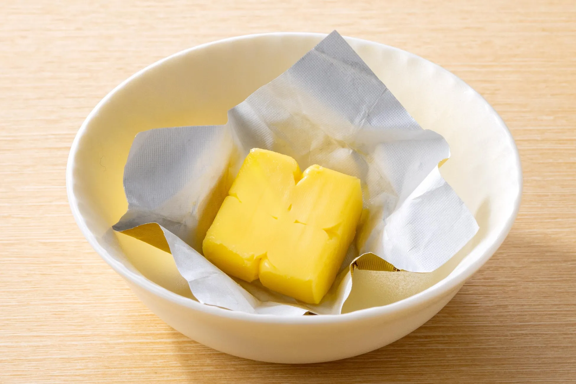 バターの塊が銀色の包装紙に包まれ、白い器に入っている様子を写した写真です。バターは黄色く、器はクリーム色で、背景は木目調のテーブルです。