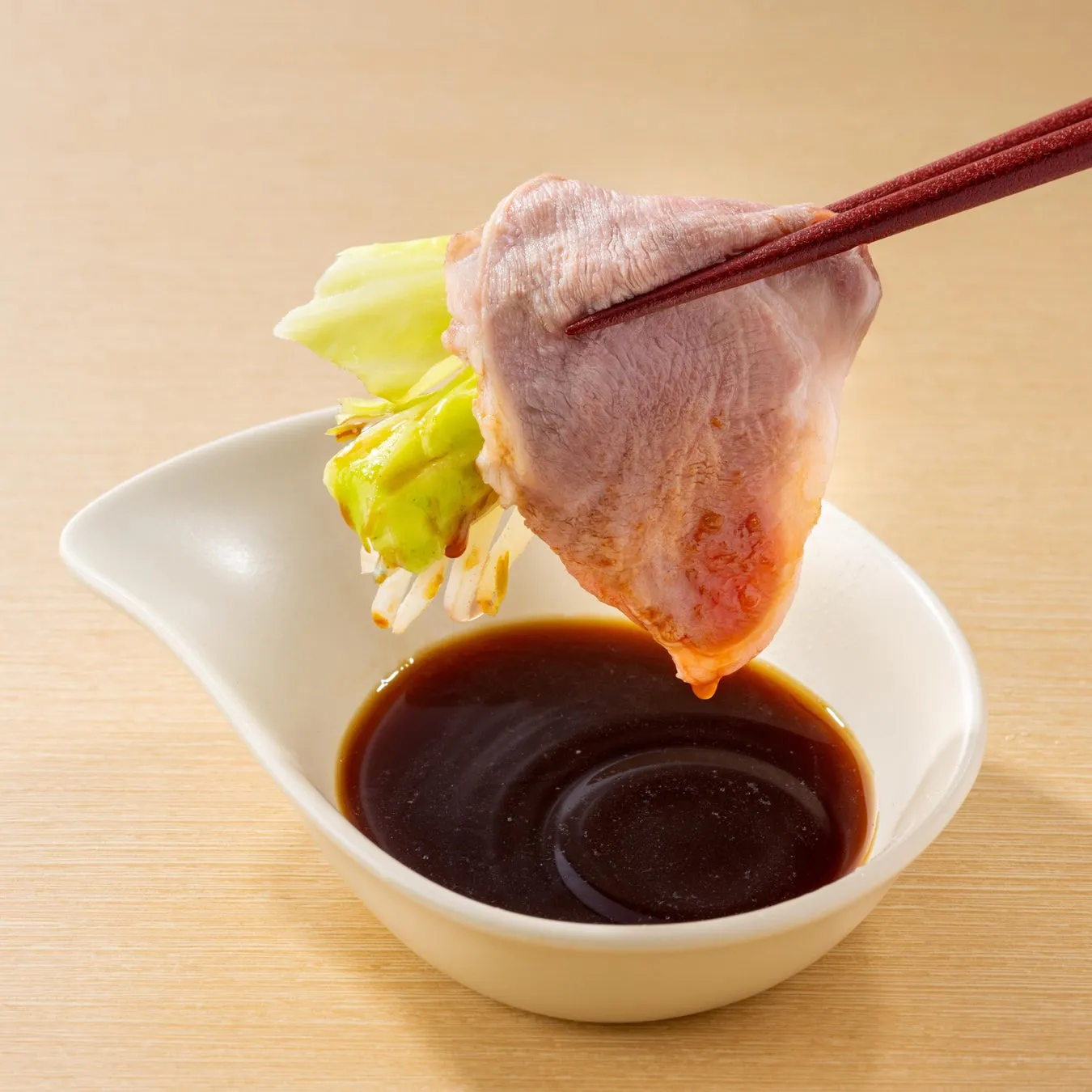 箸で持たれた豚肉と野菜が、ソースの入った器の上に配置されている様子。