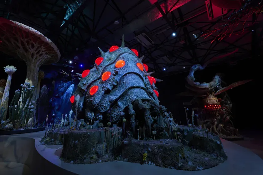 暗い空間に展示されている、巨大な生物の彫刻とキノコのような造形物。赤く光る目のようなものが特徴的。