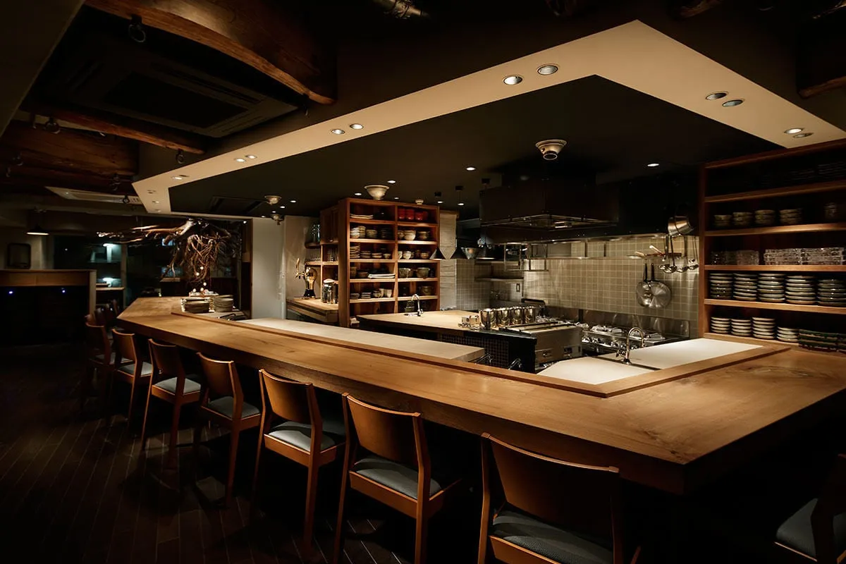 木製のカウンターと椅子が印象的な、落ち着いた雰囲気のレストランのインテリア。奥には調理場が見え、洗練された空間が広がる。