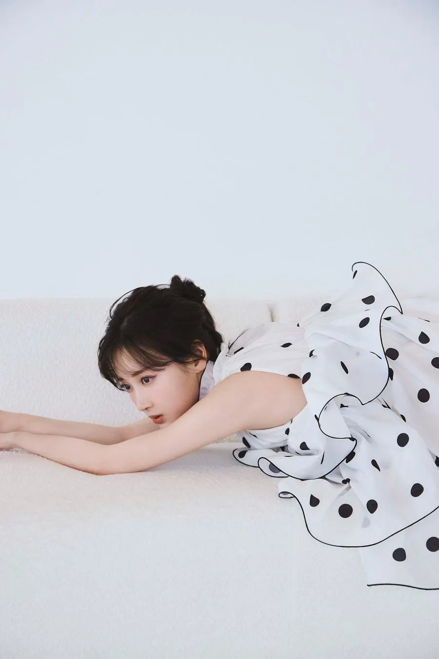 白いソファにうつ伏せで寄り添う女性。彼女は白い水玉模様のドレスを着ており、髪はまとめられています。