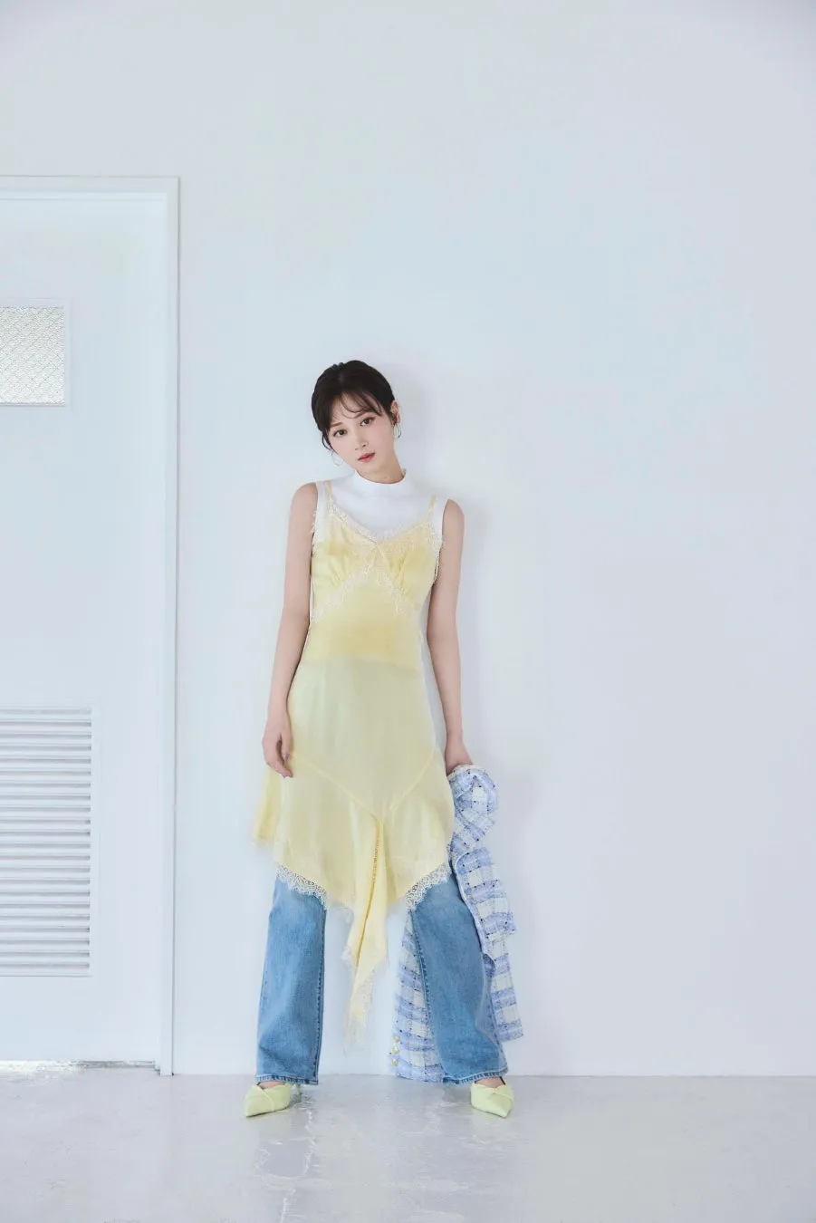 白い壁の前に立つ女性。彼女は黄色いドレスとジーンズを着ており、ジャケットを持っています。